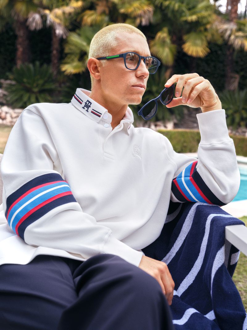 Man wearing glasses sits outside in white logo-embroidered rugby shirt and dark pants, holding sunglasses, styled by Tommy Hilfiger.