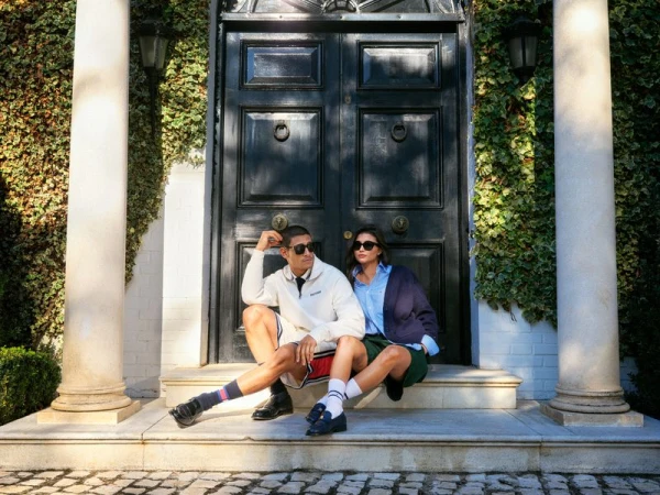 A fashionable couple poses on the front steps of a house, with dark wooden doors, columns, and ivy-covered walls in the background.