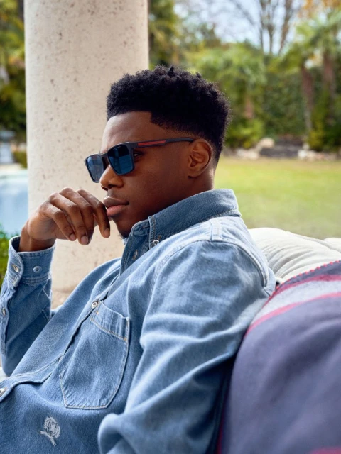 A young Black man sits outside wearing sunglasses and a denim shirt. He has a contemplative expression with green foliage in the background.