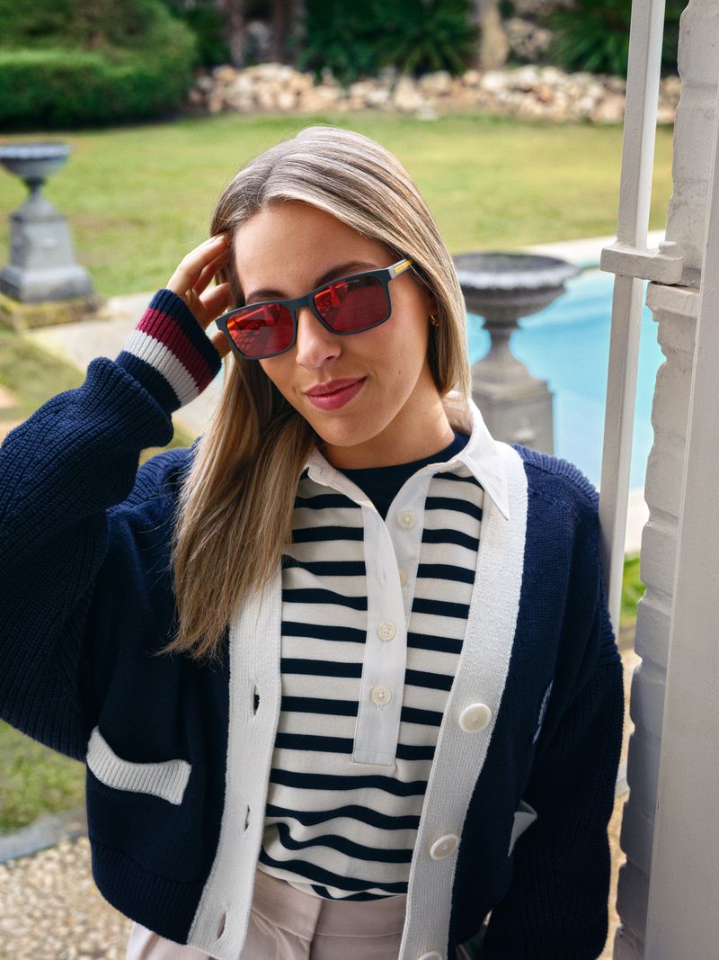 Stylish woman with blonde hair wears sunglasses and a striped shirt with a navy cardigan, posing outdoors near a pool and foliage.