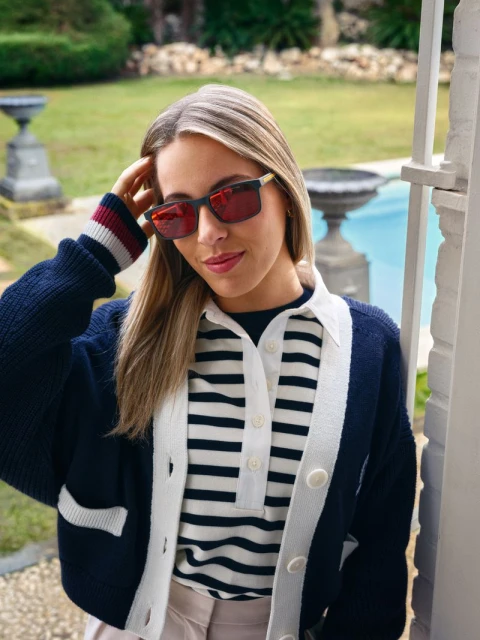 Stylish woman with blonde hair wears sunglasses and a striped shirt with a navy cardigan, posing outdoors near a pool and foliage.