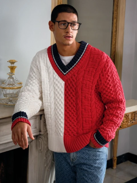 Man wearing a white and red color-blocked V-neck sweater and jeans indoors with a clear decorative glass object and white fireplace.