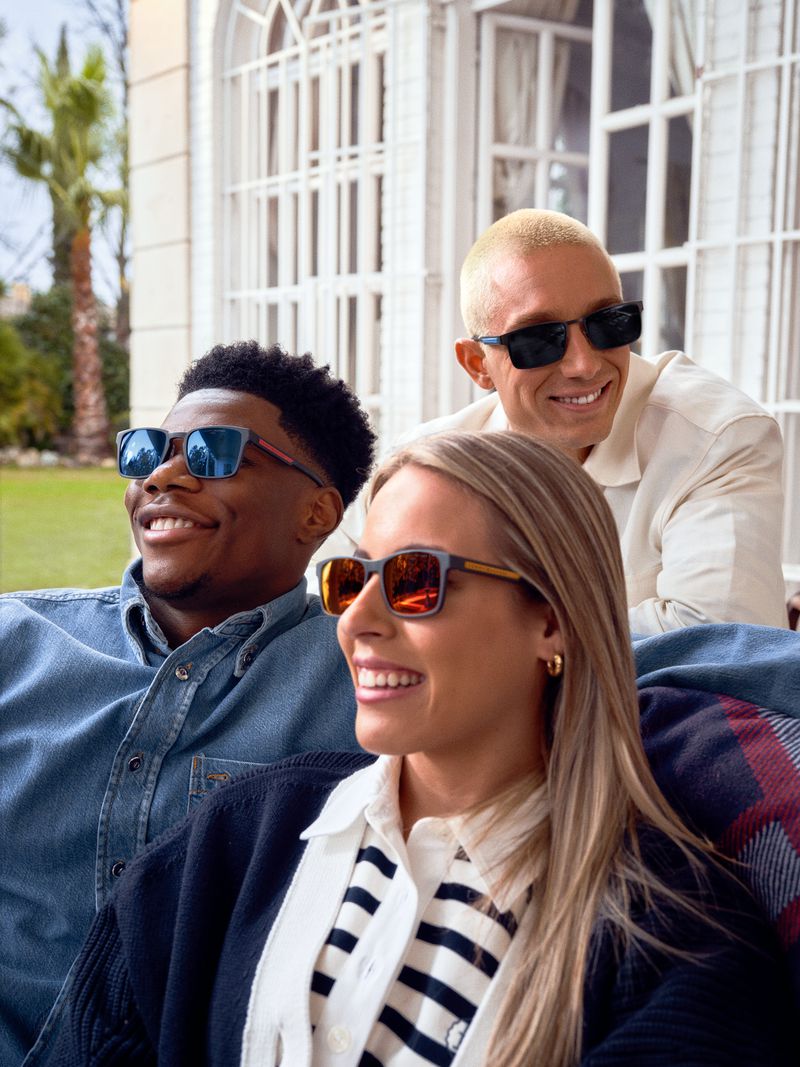 Three smiling adults wearing sunglasses outdoors. A woman in the foreground is flanked by two men who are wearing casual tops.