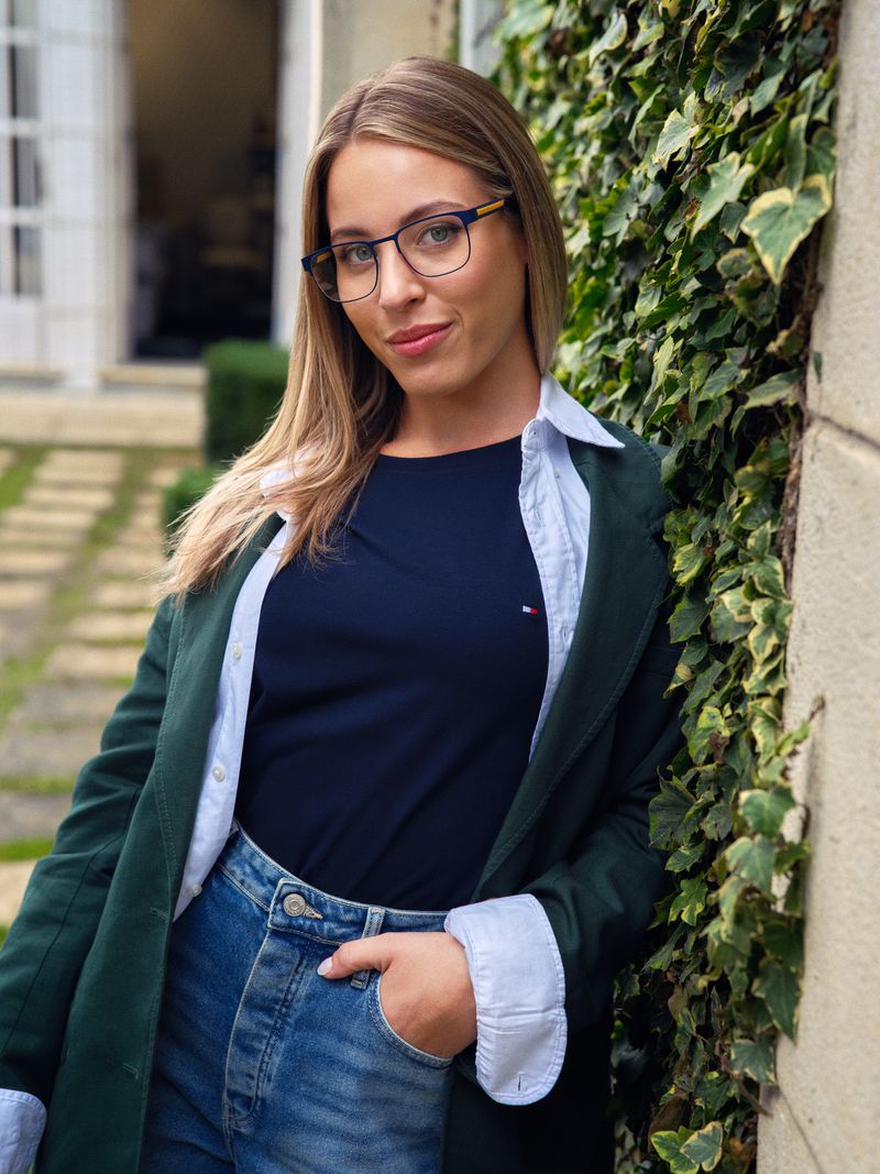 Blonde woman in eyeglasses, dark t-shirt, jeans, and green blazer stands against ivy-covered wall outdoors with blurred background buildings.