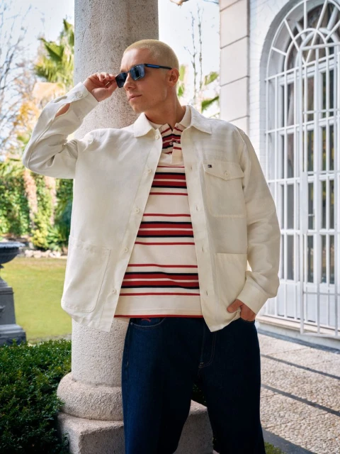 Man in unbuttoned shirt jacket over striped polo shirt and dark jeans, wearing sunglasses, poses near stone column outdoors.