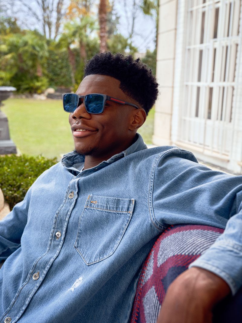 An African American man with sunglasses in a blue denim shirt smiles while sitting outdoors, backed by lush greenery and architecture.