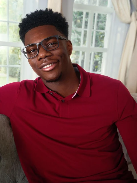 Portrait of an African-American man wearing eyeglasses and a red polo shirt sitting indoors near a window with natural light.