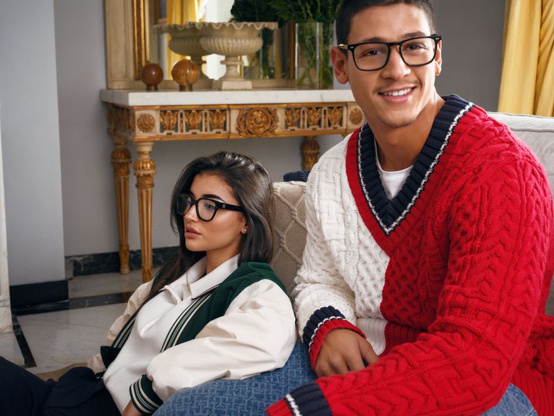 Portrait of a young couple wearing preppy fashion and eyeglasses sitting indoors; the man is wearing a red and white sweater.