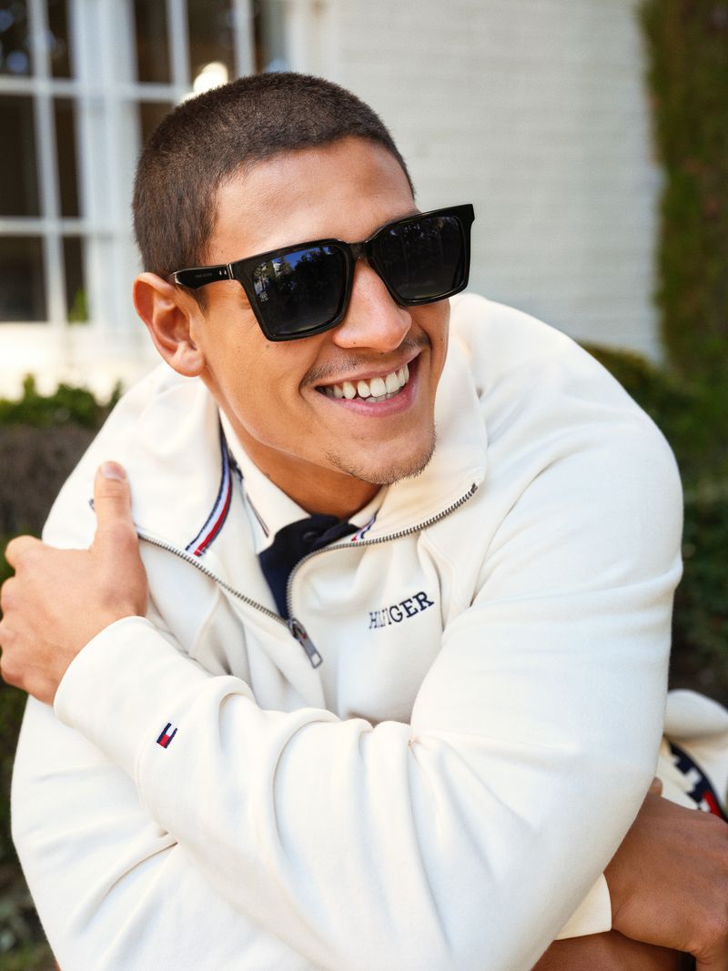 A smiling adult male with sunglasses and a jacket poses outdoors with green foliage visible in the background during daytime.