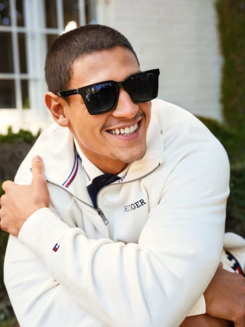 A smiling adult male with sunglasses and a jacket poses outdoors with green foliage visible in the background during daytime.