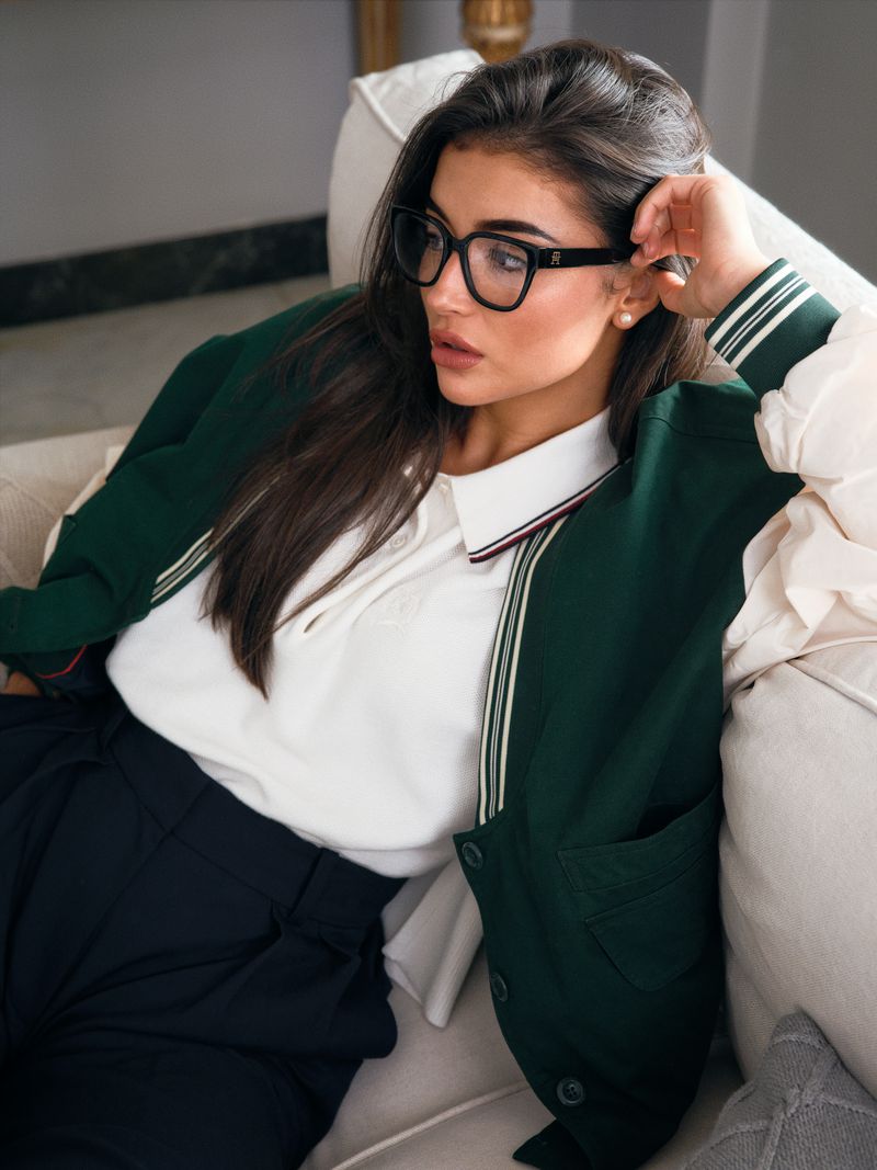 Brunette woman in glasses and green varsity jacket lounges on a cream sofa in natural light, posing elegantly for an indoor fashion shot.