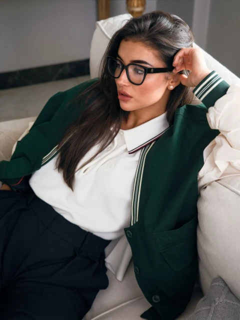 Brunette woman in glasses and green varsity jacket lounges on a cream sofa in natural light, posing elegantly for an indoor fashion shot.