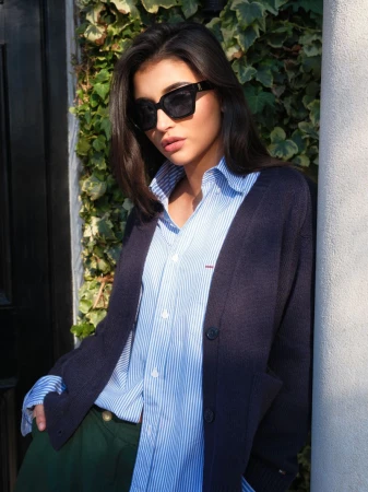 Woman wearing dark sunglasses, striped shirt, and dark blue cardigan leaning against a column outdoors.