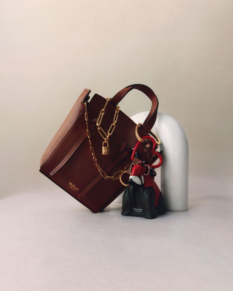 Studio shot of a brown leather Mulberry handbag accessorized with a chain and bag charm, placed against a neutral backdrop.