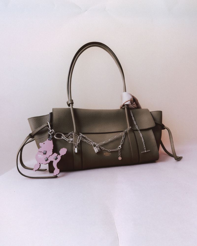 Elegant olive-green leather handbag with top handles, silver chain detail, and a pink poodle charm, presented on a white background.