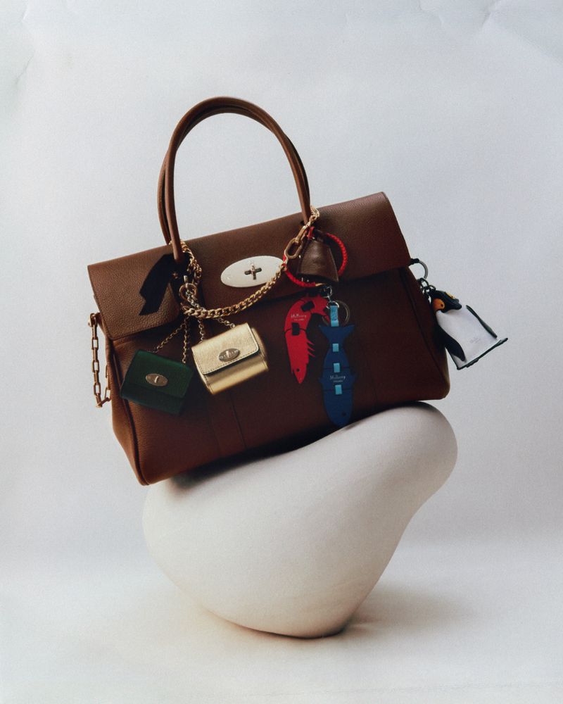 Studio shot of a brown leather handbag on a white abstract display stand, featuring attached mini bags and colorful keychains.