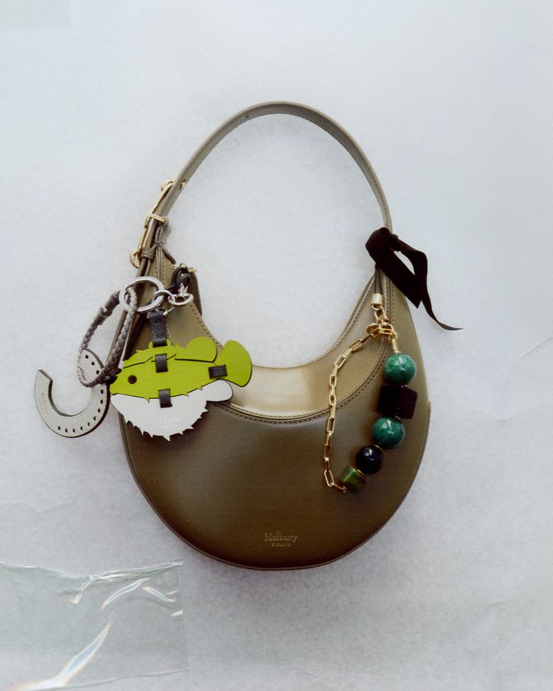 Olive green leather purse on a white surface. The purse has a rounded shape and a short strap. Gold hardware and charms dangle.