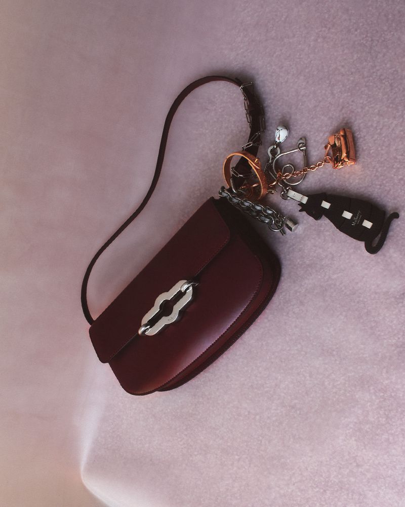 Studio shot of a dark red handbag with a shoulder strap and silver hardware, displayed on a textured light purple background.
