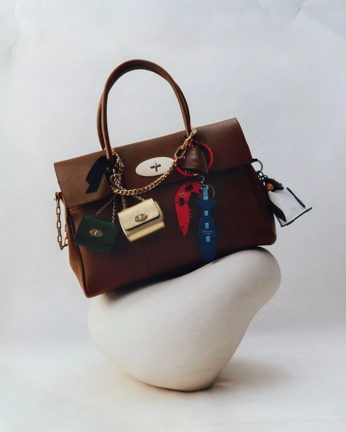Brown leather handbag decorated with multiple charms resting on a white sculptural form against a white background