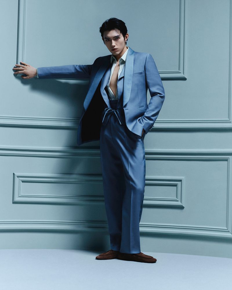 Portrait of a slender adult male in a blue suit standing in a corner against a blue-panelled wall inside a photo studio.