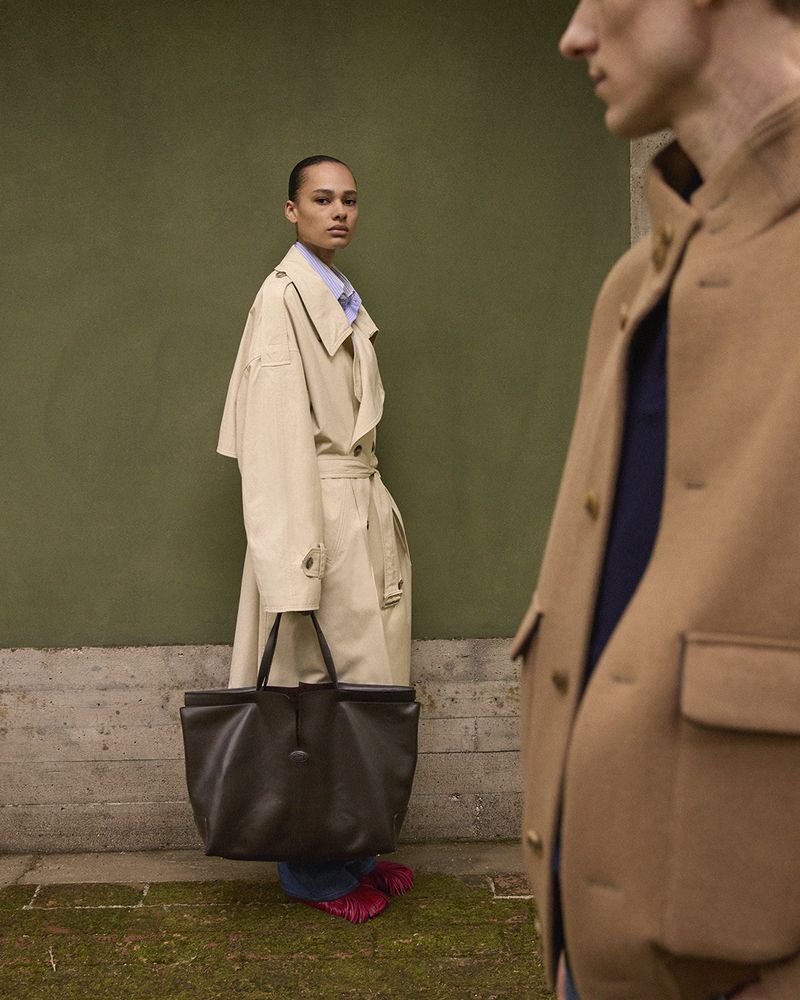 Two people in tan coats stand against a green wall. A woman with a large black leather bag is on the left. Fashion photography.