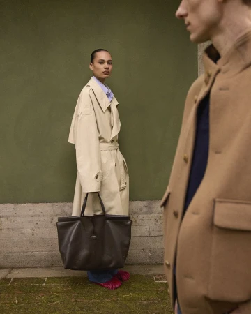 Two people in tan coats stand against a green wall. A woman with a large black leather bag is on the left. Fashion photography.