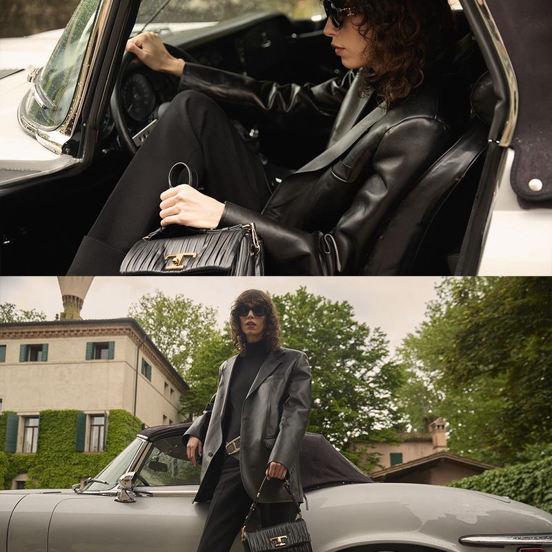 An adult female model in a black leather jacket is driving a convertible car in an outdoor setting on a cloudy day.