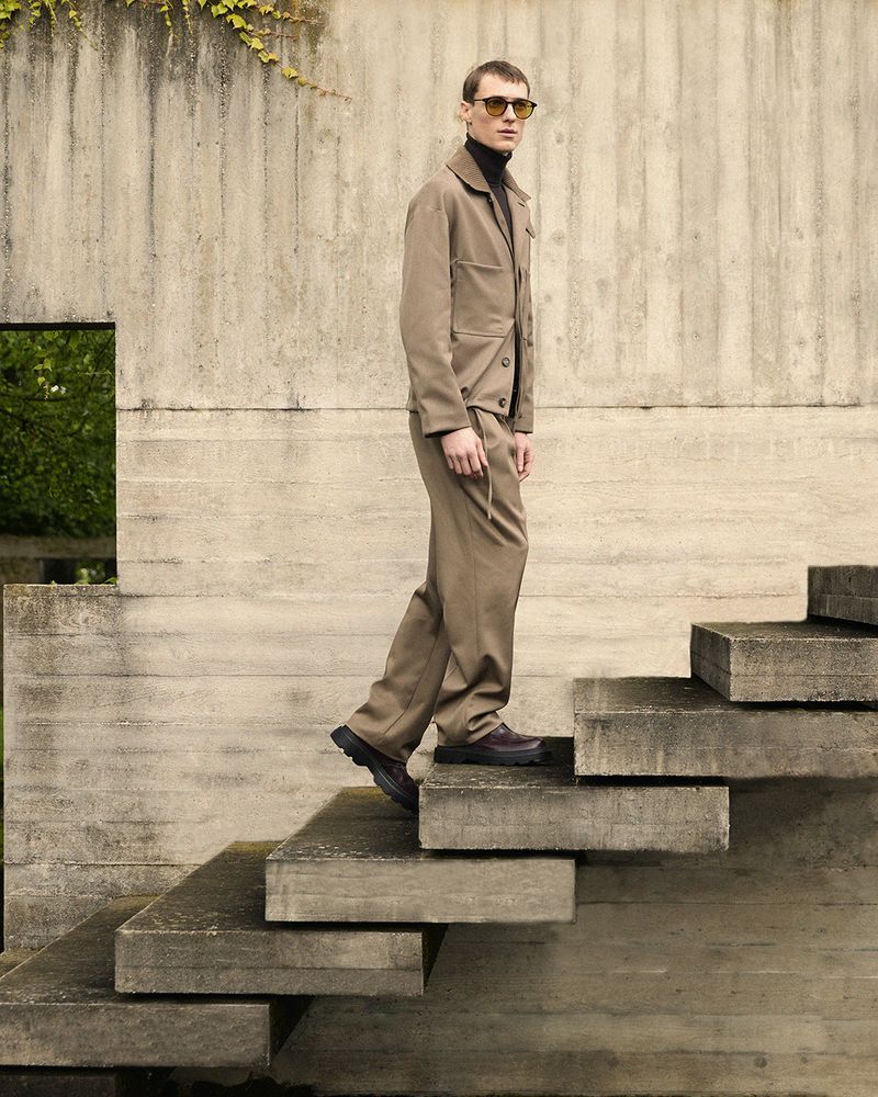 An adult male in a tan suit ascends concrete stairs outdoors, showcasing fashion and architectural elements in natural light.