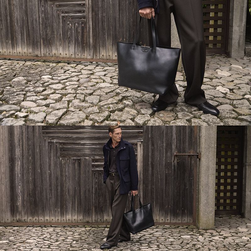 Man in blue jacket and brown pants holds a black tote bag on a cobblestone street near a weathered wooden gate and stone wall.