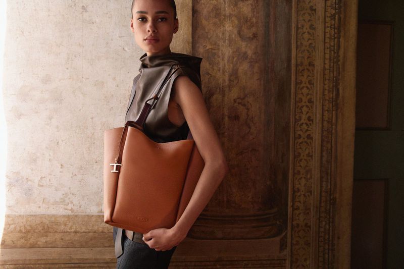 A woman in a leather outfit poses indoors, holding a brown leather tote bag with a 'T' charm. Fashion, style, and luxury are highlighted.