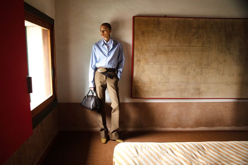 A stylishly dressed adult female stands in a light-filled room with artwork, modelling a light-blue button-down shirt and brown slacks.