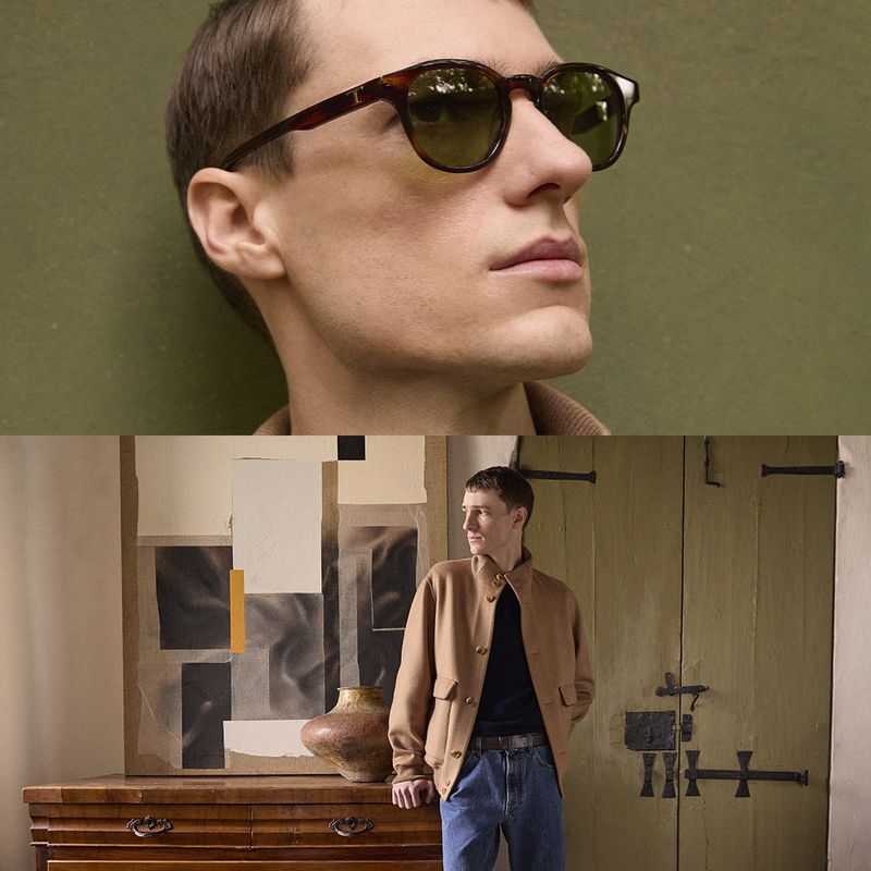 Man wearing sunglasses and a tan jacket poses indoors with artwork and a vintage door, showcasing a blend of classic and modern styles.