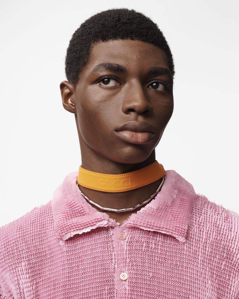 Close-up of a dark-skinned male model in a pink textured shirt wearing an orange choker with visible 'GUCCI' text against a white backdrop.