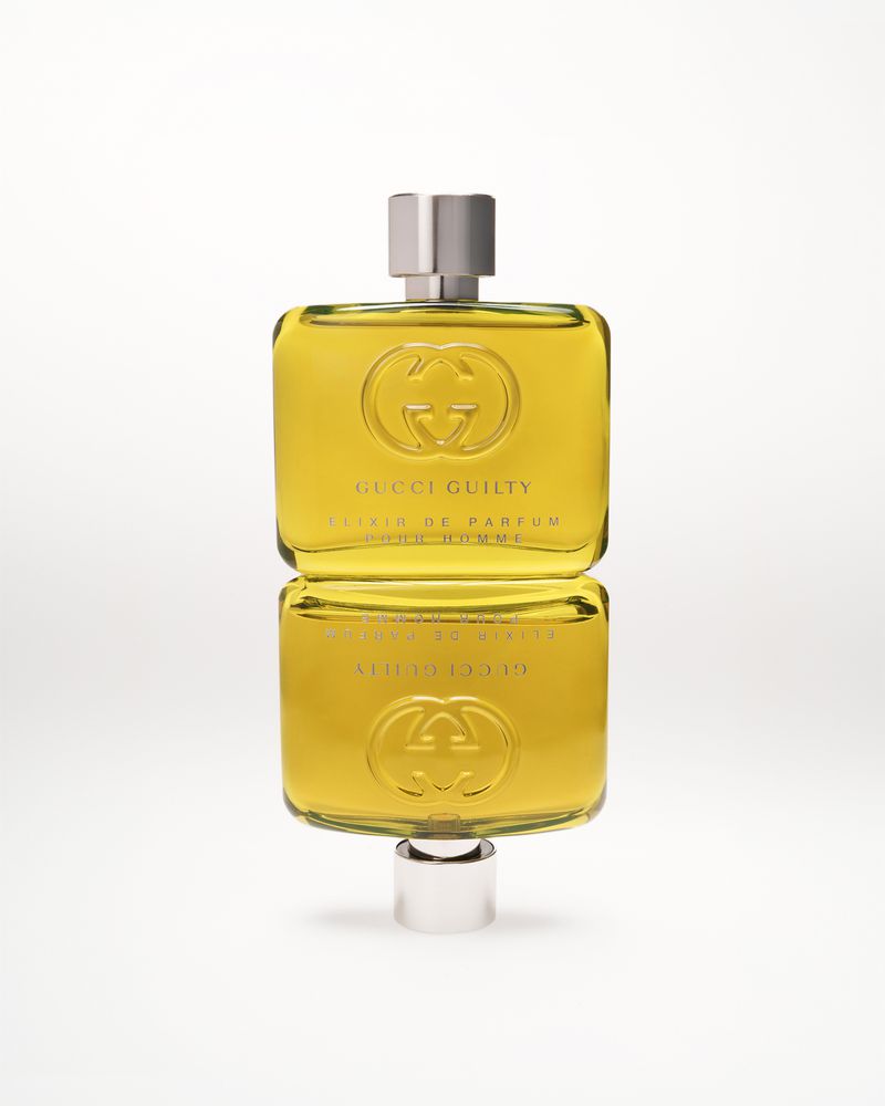 Gucci Guilty Elixir de Parfum bottle reflecting on a white surface. Silver cap and gold liquid give a luxurious feel.