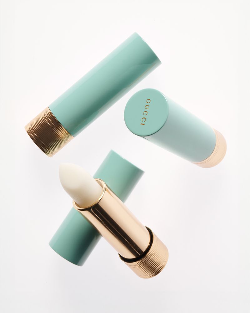 Three tubes of Gucci Rouge de Beauté lip balm in light blue and gold tones are featured against a white background in a studio shot.