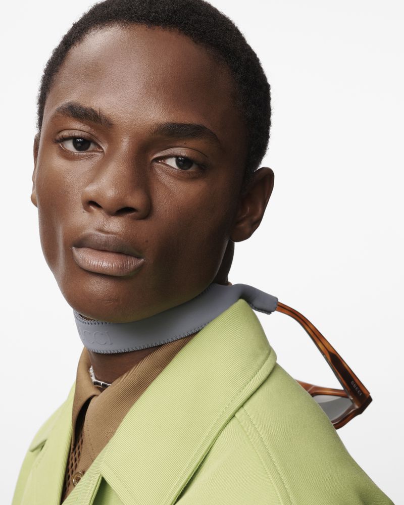 Close-up portrait of a stylish African-American man wearing a lime green jacket, glasses, and a choker necklace against a white background.