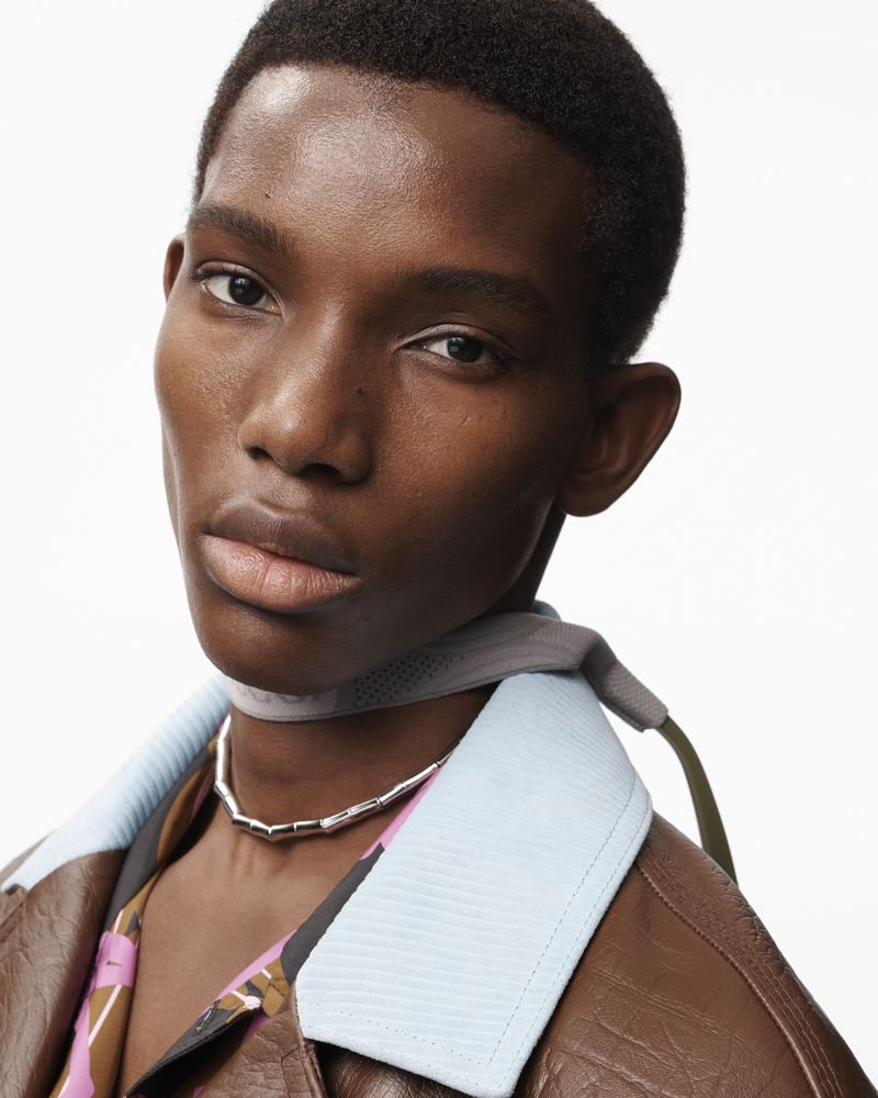 Close-up of a black male model with short hair wearing a brown jacket, silver necklace, and patterned shirt against a white background.