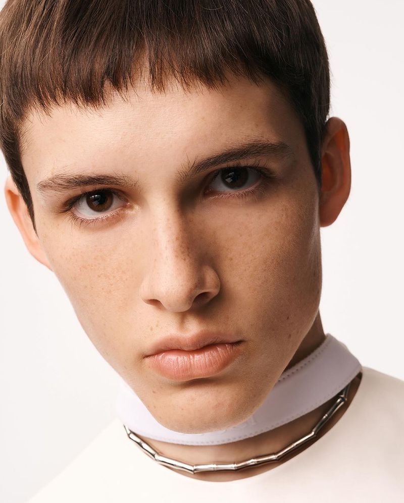 Close-up studio shot of an adult male with a neutral facial expression, wearing a cream-colored top and a silver necklace.