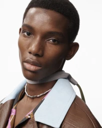 Close-up portrait of a male model wearing a brown jacket with a pale blue collar and a silver necklace against a white background.