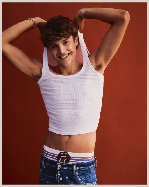 Young man smiles, pulling up white tank top, showcasing branded waistband and jeans against a solid burnt orange background in studio lighting.