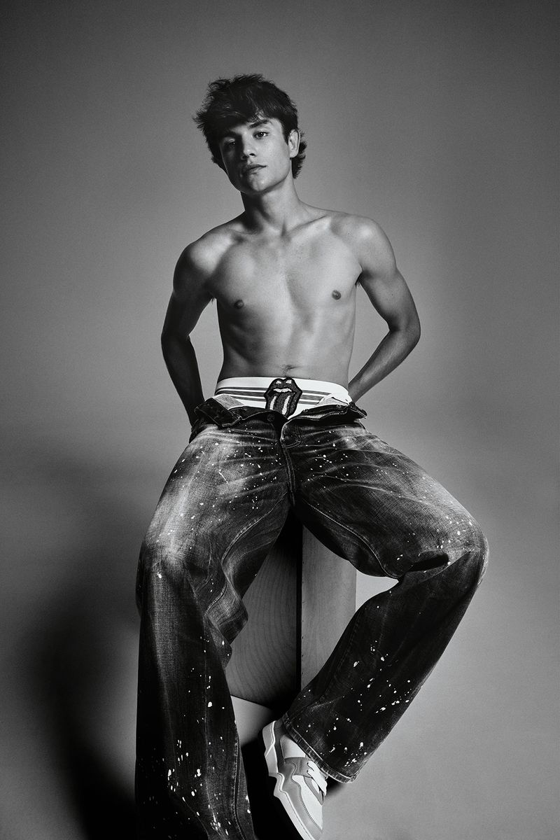 Black and white portrait of a shirtless young man in jeans and sneakers sitting on a wooden block in a photo studio.