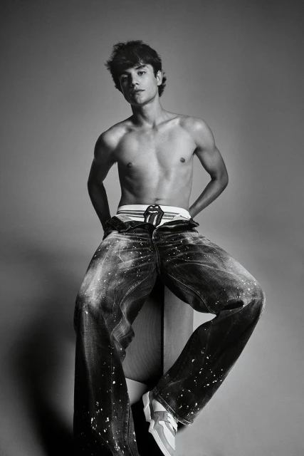 Black and white portrait of a shirtless young man in jeans and sneakers sitting on a wooden block in a photo studio.