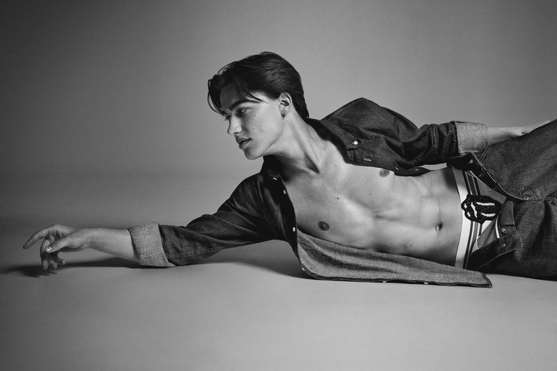 Monochrome studio shot of a reclining adult male fashion model wearing an unbuttoned denim shirt and white boxer briefs with black stripes.