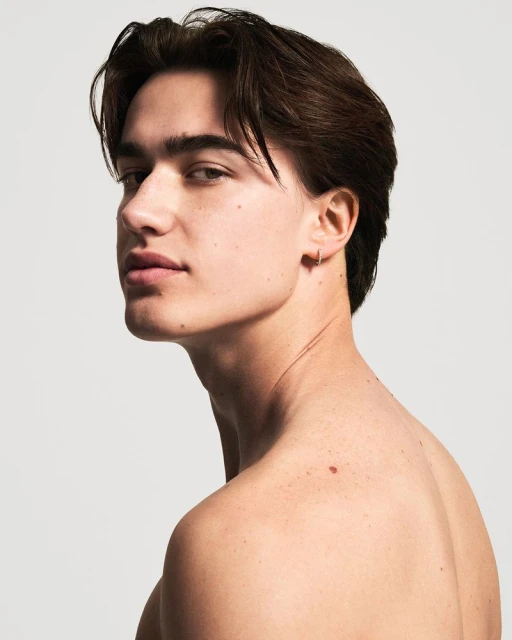Close-up studio portrait of a young adult man with dark brown hair, fair skin, earring, mole, posing against a neutral light gray background.
