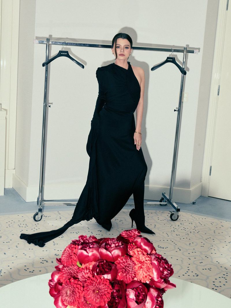 Elegant woman in a black gown poses in a studio with a rack of clothes behind her and a red flower arrangement in the foreground.