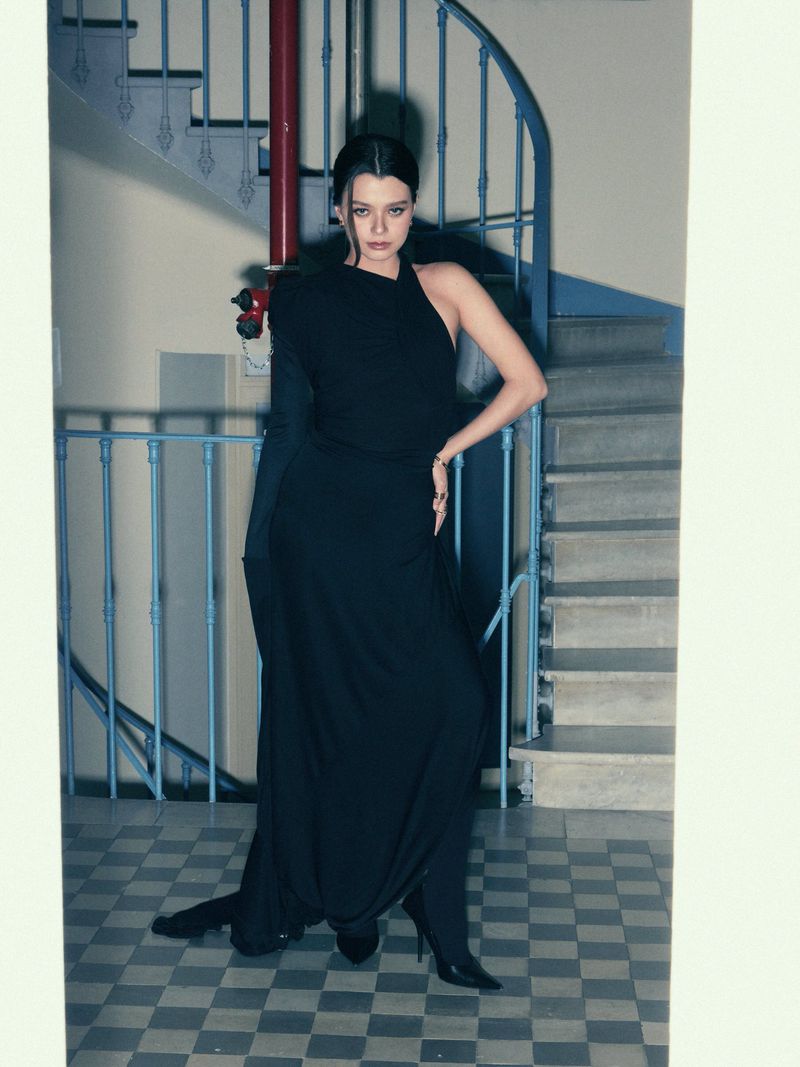 A brunette woman poses in an elegant black dress against a tiled floor and spiral staircase, showcasing modern fashion in an indoor setting.