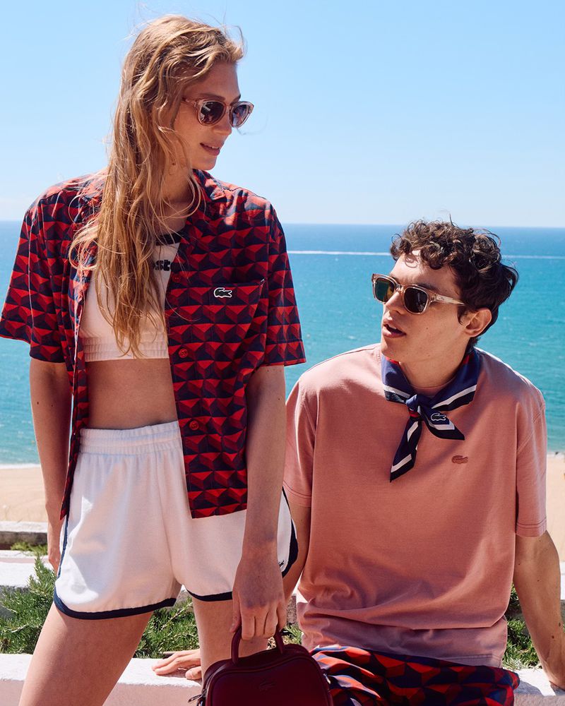 Two young adults pose with Lacoste apparel near a scenic beach; a woman in a patterned shirt holds a small bag, and a man wears a t-shirt.