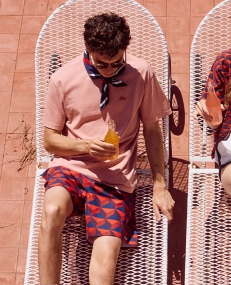 Man lounges in a white metal chair with geometric-patterned shorts, pink t-shirt, and sunglasses, holding a glass bottle drink on a rooftop.