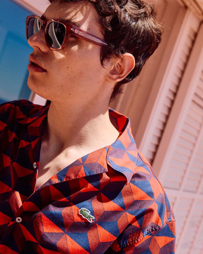 Young man wearing sunglasses and a patterned Lacoste shirt, posing outdoors with sunlight and geometric shapes in the background.