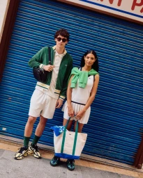 Two models standing in front of a blue roller shutter door wearing Lacoste sportswear, shorts, and holding bags.